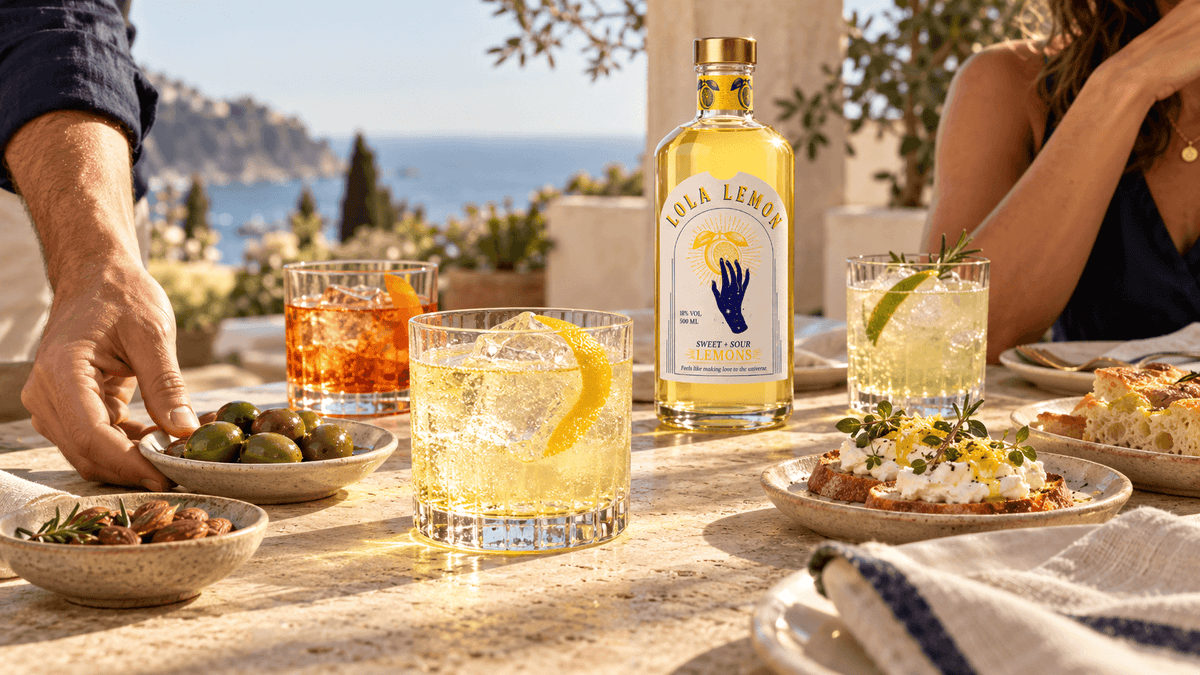 Drei Aperitifgläser und eine Flasche LOLA Lemon mit Oliven, Mandeln und Crostini auf einer sonnigen Terrasse mit Meerblick.