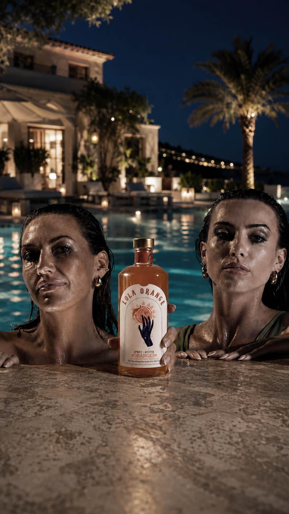 Zwei Frauen im nächtlich beleuchteten Pool präsentieren eine Flasche LOLA ORANGE Aperitif in eleganter Sommeratmosphäre.