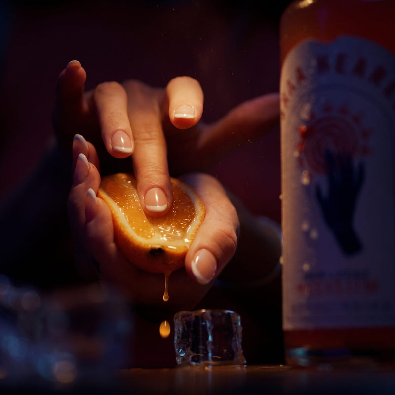 Sommerlicher LOLA ORANGE Moment: Eine Frau genießt einen Aperitif-Drink, während eine frische Orange neben der LOLA ORANGE Flasche ausgedrückt wird.