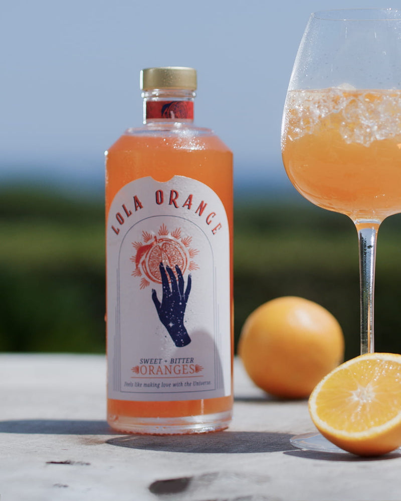 LOLA ORANGE Aperitif-Flasche mit eisgekühltem Orange-Drink und frischen Orangen in sommerlicher Atmosphäre.
